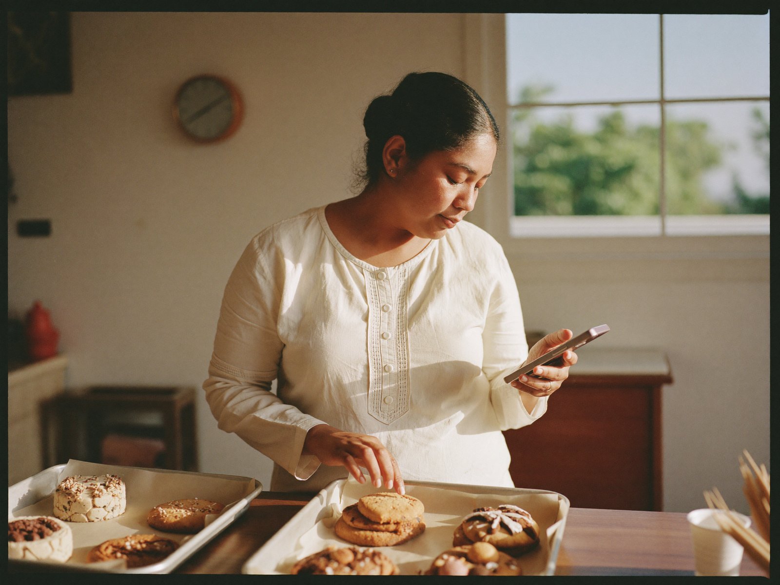 A home bakery owner checking her phone beside trays of freshly baked cakes and cookies.